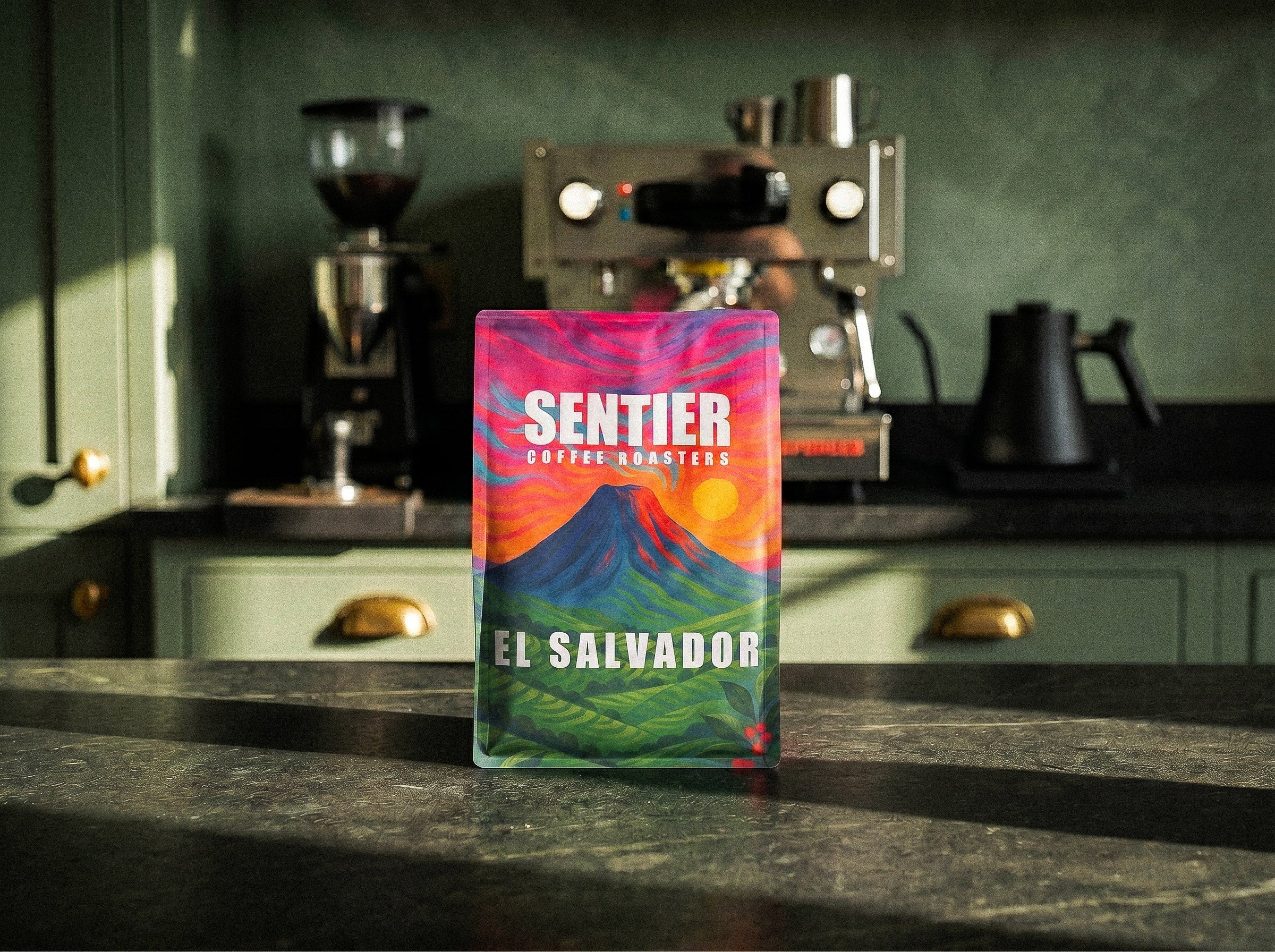 Colorful coffee bag labeled 'Sentier Coffee Roasters' on a kitchen counter with coffee-making equipment in the background.