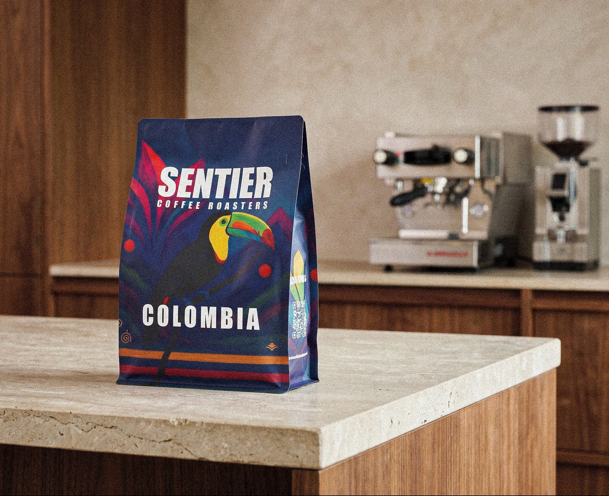 Sentier Coffee Roasters Colombia coffee bag on a kitchen counter with espresso machines in the background.